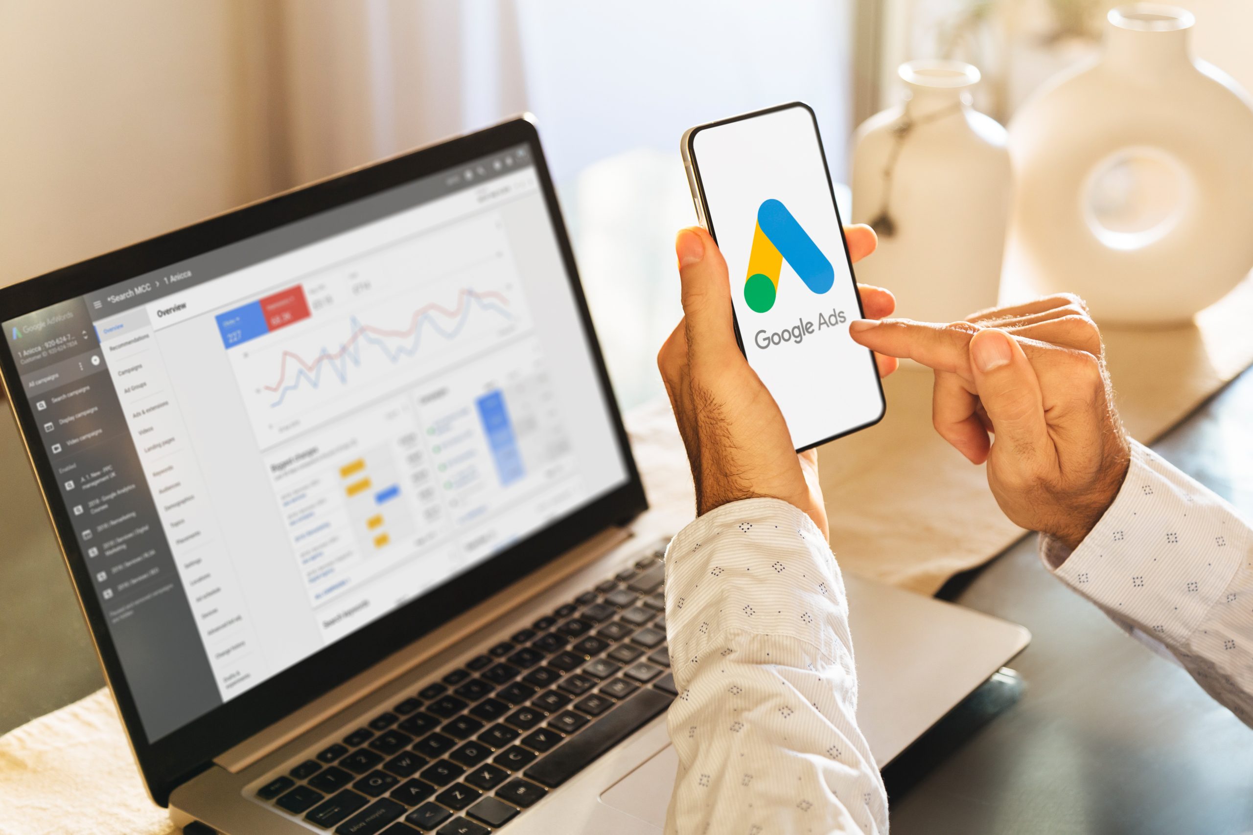Man using a mobile phone with Google Ads on the screen and in the background a computer showing ad statistics.